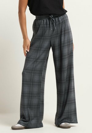 Osoba ubrana w szerokie szare spodnie w kratę z elastycznym pasem i sznurkiem, w połączeniu z białymi i szarymi trampkami.