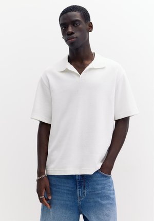 Mladý muž stojaci s rukami v vreckách, oblečený v bielej polo košeli s krátkym rukávom a modrých džínsoch, na jednoduchom bielom pozadí.