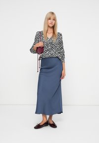 Blouse à motif zèbre noir et blanc, jupe maxi en satin bleu marine et chaussures plates bordeaux. Les accessoires incluent un petit sac croisé bordeaux.