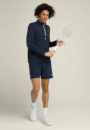 Navy athletic top with a half-zip design and white accents, paired with matching shorts. Holding a white tennis racket with red detailing.