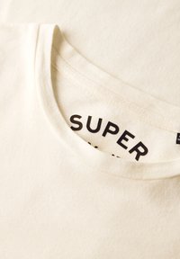 Crème-kleurige katoenen t-shirt met een ronde halslijn; heeft zwarte tekst binnen de kraag met de tekst "SUPER" en een zachte, gladde textuur.