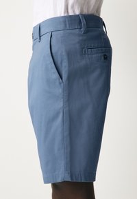 Svetlo modre bombažne kratke hlače z ravnim sprednjim delom, stranskimi žepi in zadnjim žepom z odprtino. Imajo zapenjanje na gumb in zanke za pas.