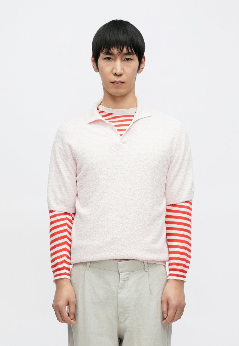 Ροζ ανοιχτόχρωμο πλεκτό πουλόβερ με κοντά μανίκια, φορεμένο πάνω από κόκκινο και λευκό ριγέ πουκάμισο με μακριά μανίκια, συνδυασμένο με ανοιχτό γκρι παντελόνι.