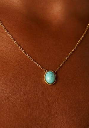 Gouden ketting met een ovale turkooizen steen met dunne zwarte aderen, rustend op een bruine huid.