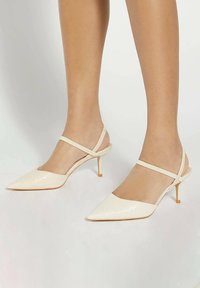 Escarpins en cuir verni beige à talon aiguille et bout pointu. Présentent des brides chevilles croisées et une finition texturée.