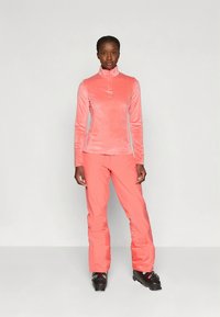 Világos rózsaszín polár pulóver magas nyakkal és fél cipzárral, hozzáillő rózsaszín sí nadrággal és fekete sícipővel párosítva.