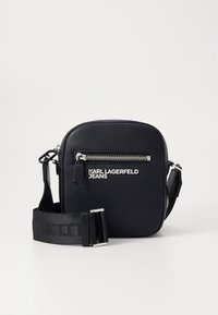 Črna usnjena torbica čez ramo s srebrnim zadrgo in širokim nastavljivim trakom, z blagovno znamko "Karl Lagerfeld Jeans" na sprednjem �žepu.