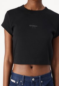 Melna īsā T-krekls, izgatavots no ribota auduma, ar apaļu kakla izgriezumu un īsām piedurknēm, centrā ar smalku Calvin Klein logo.