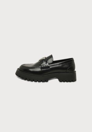 Mocassin en cuir noir avec semelle extérieure à crampons épaisse et détail boucle en métal sur un fond clair uni.