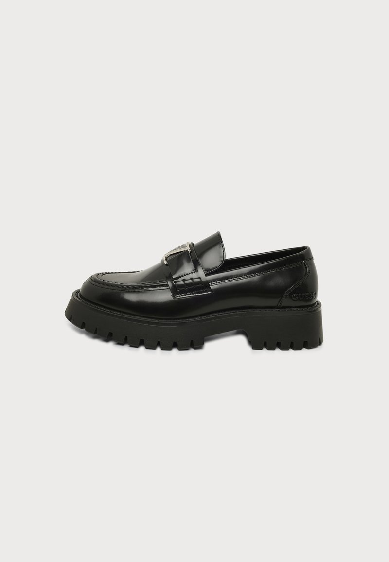 Mocassin en cuir noir avec semelle extérieure à crampons épaisse et détail boucle en métal sur un fond clair uni.
