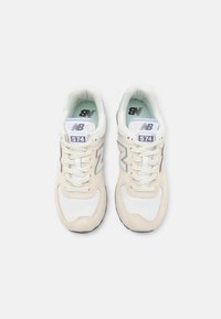Beige und weiße New Balance 574 Sneaker mit Obermaterial aus Wildleder und Textil, reflektierenden Akzenten, Textilauskleidung und marmorierter Gummisohle.