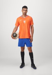 Oranje Nike-shirt met een gestructureerd patroon, witte kraag en logo, gecombineerd met blauwe shorts, zwarte sokken en zwarte voetbalschoenen, terwijl hij een voetbal vasthoudt.