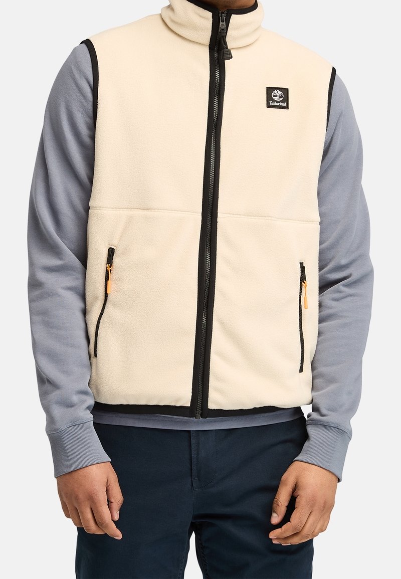 Homme portant un gilet polaire beige sans manches avec une fermeture éclair noire et un écusson logo, par-dessus une chemise grise à manches longues et un pantalon foncé.