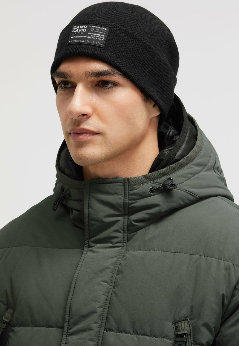 Olive green puffer jacket with a high collar and black zippered pockets. Black knitted beanie features a label on the front.