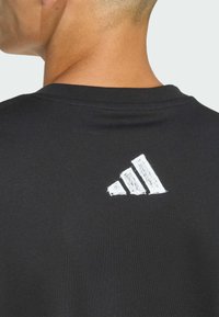 Schwarzes Kurzarmshirt mit einem weißen, strukturierten Drei-Streifen-Logo auf dem Rücken. Hergestellt aus glattem Stoff mit Rundhalsausschnitt.