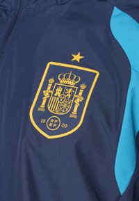 Navy athletic jacket with light blue accents, featuring a gold shield emblem with a crown, symbols, and the year 1909. Smooth fabric texture.