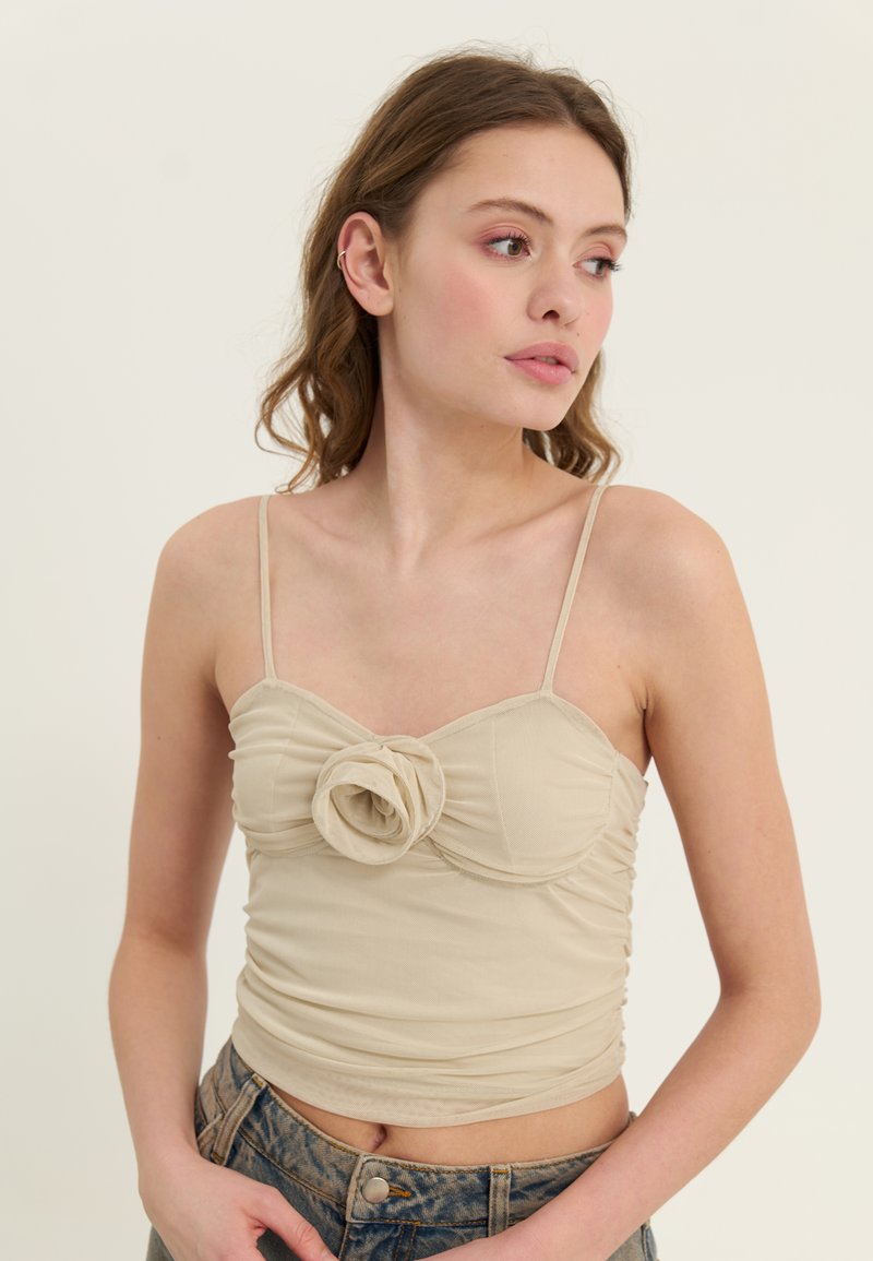 Beige crop top med tynne stropper, en rynket design, og en dekorativ stoffblomst i midten. Brukt sammen med høyhalsede jeans.