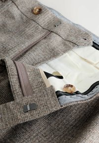 Braune Tweed-Hosen mit Reißverschluss, Innenfutter in Creme und braunen Knöpfen; mit einer schwarz-weißen gemusterten Borte.