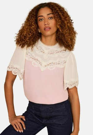 Femme aux cheveux bouclés portant un chemisier en dentelle blanche à manches bouffantes et un jean foncé taille haute, posant avec une main sur la hanche.