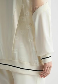 Bílá průsvitná blůza s logem; má kontrastní černý pruh na man�žetách a spodním lemu, s jemnou, lehkou texturou.