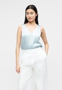 Γυναίκα με μακριά μαύρα μαλλιά που φοράει ανοιχτό μπλε αμάνικη σατέν μπλούζα μέσα σε λευκό, φαρδύ παντελόνι με ψηλή μέση, στέκεται με το ένα χέρι στην τσέπη.