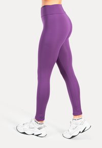 Paarse sportlegging met een hoge tailleband, een gladde textuur en een nauwsluitend ontwerp. Draagt witte sneakers met zwarte accenten.