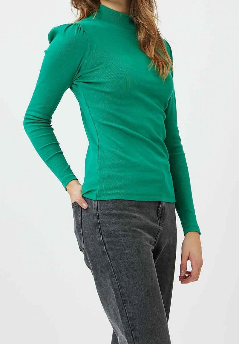 Grön långärmad topp med hög krage och rynkad axeldetalj, tillsammans med mörkgrå byxor. Texturerat, figursytt tyg.