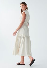 Kreemja värvi keskmise pikkusega kleit lühikeste varrukatega, rõhutatud talje ja kihilise äärisega. Mudel kannab musti sandaale. Lihtne tekstuur.