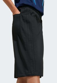 Black knee-length shorts with triple side stripes worn with a navy blue shirt, shown with a hand in the back pocket.