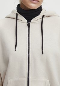 Kreminė zip-up gobtuvė su juodu užtrauktuku ir raišteliais. Savybės: minkšta tekstūra ir didelės priekinės kišenės, skirtos patogumui.