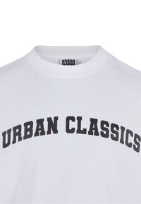 Balta medvilninė marškinėliai su apvaliu iškirpimu, ant kurių priekio didelė juoda "URBAN CLASSICS" užrašas paryškintomis, išlenktomis raidėmis.
