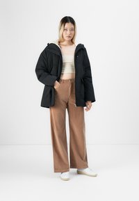 Ung kvinne står med en hånd i lommen iført svart boblejakke, beige kort topp, brede brune cordpantalon og hvite sneakers.