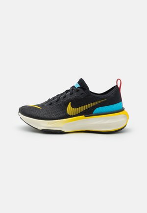 Zwart en geel Nike hardloopschoen met dikke witte zool, blauw accent bij de hiel, rood trekkoordje en geel Swoosh-logo aan de zijkant.
