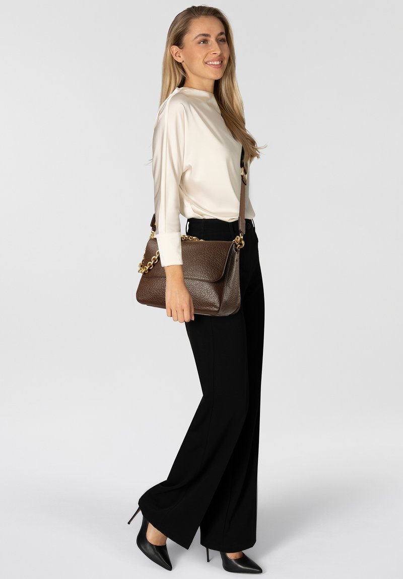 Brown textured leather handbag with gold hardware, adjustable strap, and flap closure. Model wears a long-sleeve ivory top and black pants.