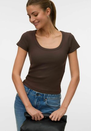 Young woman with brown scoop-neck short sleeve top and blue jeans holding a folded black leather bag in front of her waist.