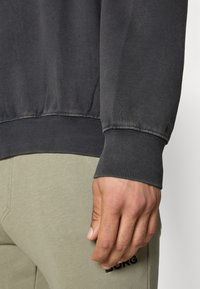 Sweatshirt cinza escuro com bainha e punhos canelados, feito de material macio. Close-up de uma mão e calças verdes com detalhe de logotipo.