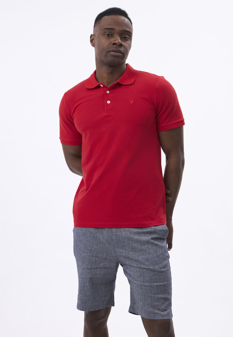 Rød polo-shirt lavet af bomuld med en tre-knaps placket, korte ærmer og et diskret logo på brystet; parret med grå shorts.