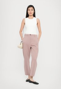 Débardeur blanc, pantalon rose sur mesure avec poches avant et ballerines noires. Le modèle tient un petit sac beige avec une chaîne.