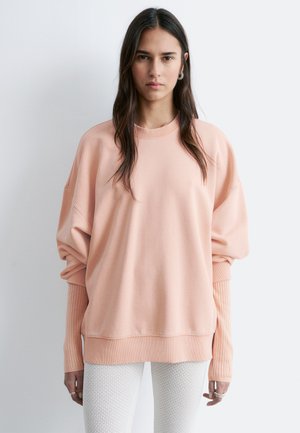 Kobieta o długich ciemnych włosach w oversize'owej morelowej bluzie z prążkowanymi długimi rękawami i białych teksturowanych spodniach, stojąca na jasnym tle.