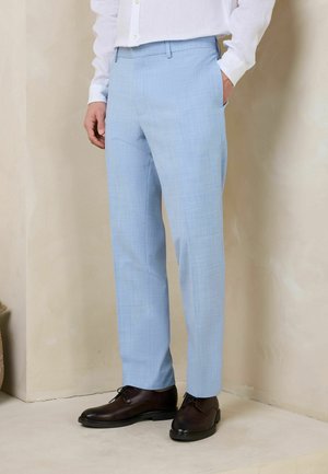 Man wearing light blue tailored trousers and dark brown leather dress shoes, standing against beige wall with one hand in pocket.