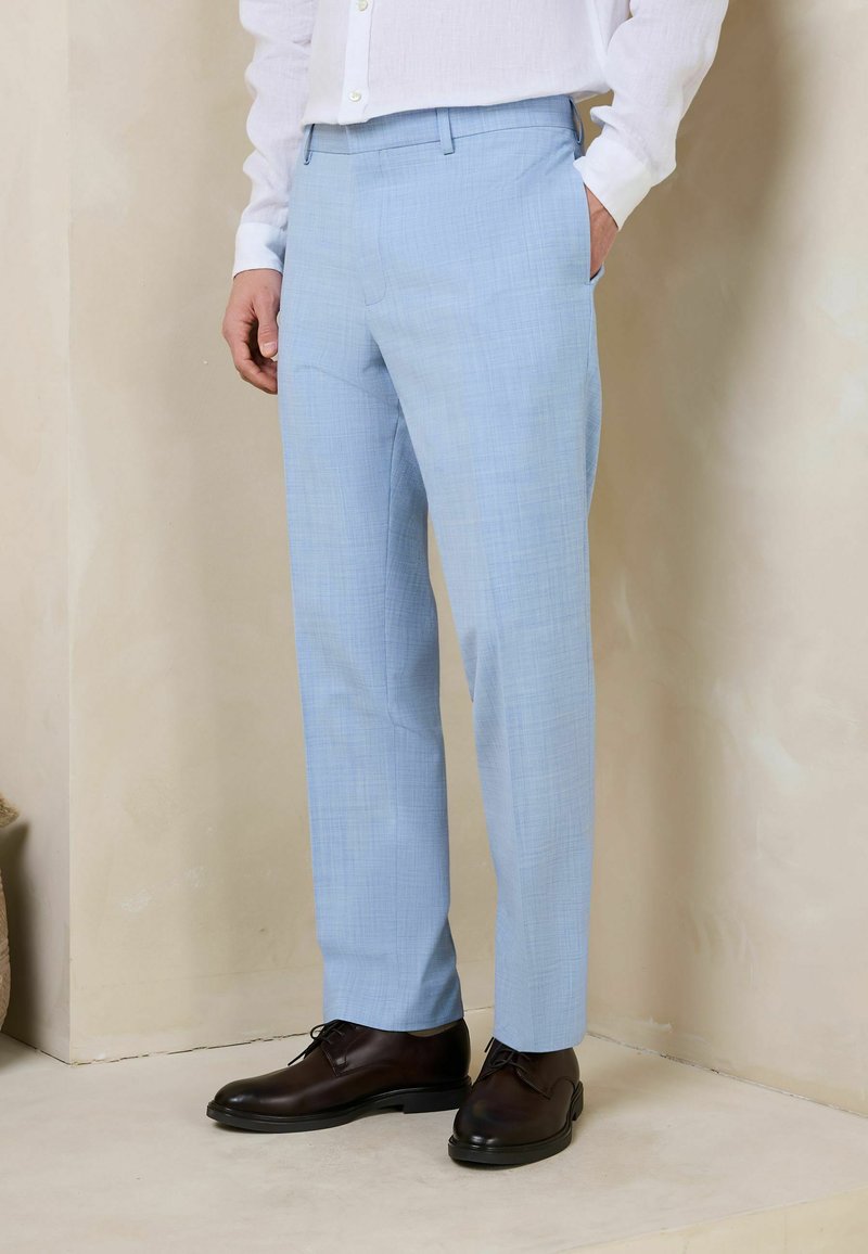 Homme portant un pantalon bleu clair sur mesure et des chaussures en cuir marron foncé, debout contre un mur beige avec une main dans la poche.