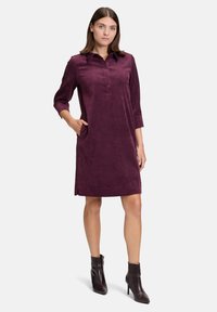 Frau steht mit der Hand in der Tasche, trägt ein knielanges lila Kleid und dunkle Knöchelstiefel vor einem einfarbigen hellen Hintergrund.