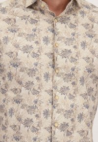 Beige overhemd met knopen, versierd met blauwe en bruine bloem- en bladvormen, gedragen door een persoon die van nek tot midden van de romp te zien is.