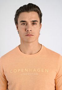 Camiseta de manga larga en color melocotón claro con cuello redondo, con "COPENHAGEN LINDBERGH" impreso en un tono más claro en la parte delantera.
