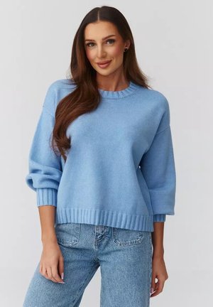 Kobieta z długimi brązowymi włosami, ubrana w jasnoniebieski sweter i niebieskie dżinsy, stojąca na tle jednolitego jasnoszarego tła.