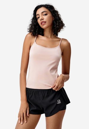 Frau mit lockigem dunklem Haar, die ein hellrosa ärmelloses Top und schwarze Sportshorts mit weißem Logo trägt, steht vor einem einfarbigen Hintergrund.
