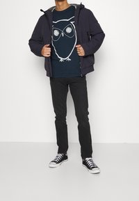 Tummansininen takki harmaalla vuorilla, yhdistettynä laivastonsiniseen T-paitaan, jossa on valkoinen pöllökuvio, sekä mustiin farkkuihin, joita komplementoi musta urheilukengät.