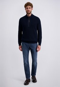 Navyblauwe gebreide pullover met een kwart-rits kraag, lange mouwen en een textuurpatroon, gecombineerd met blauwe jeans en bruine schoenen.