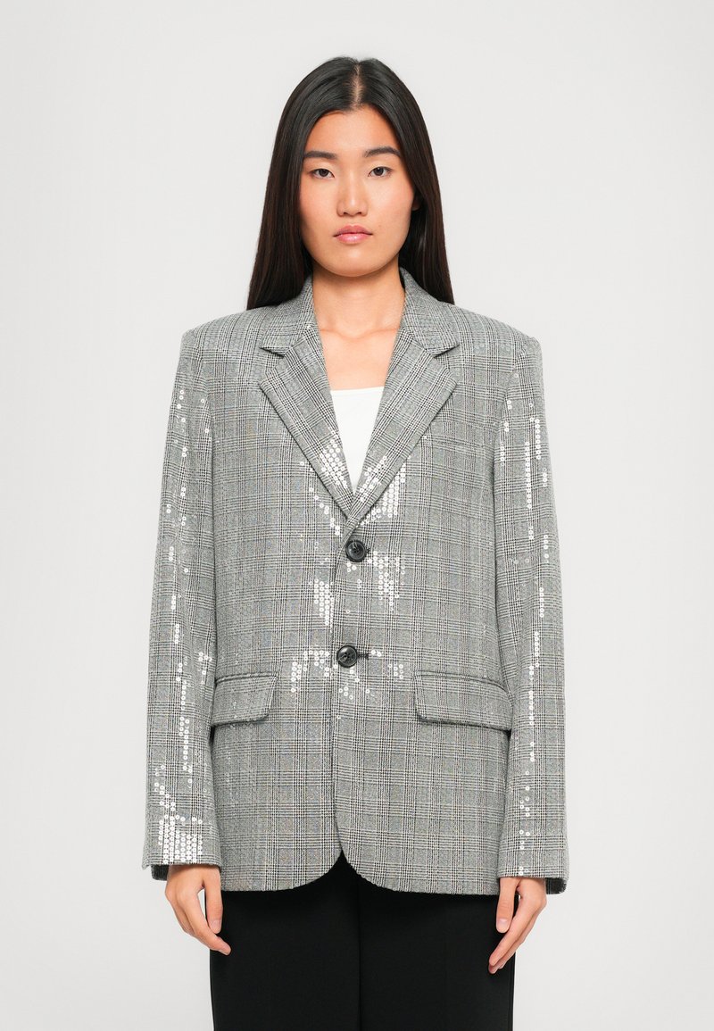 Donna con capelli lunghi e scuri, che indossa un blazer grigio a quadri con dettagli luccicanti di paillettes e pantaloni neri, su uno sfondo chiaro e semplice.