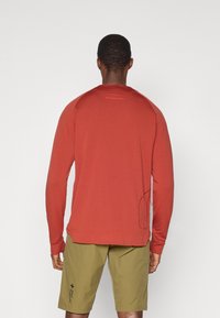 Langermet rød skjorte med teksturert stoff, rund hals, og subtile logo-detaljer på baksiden, kombinert med lyse olivengrønne shorts.